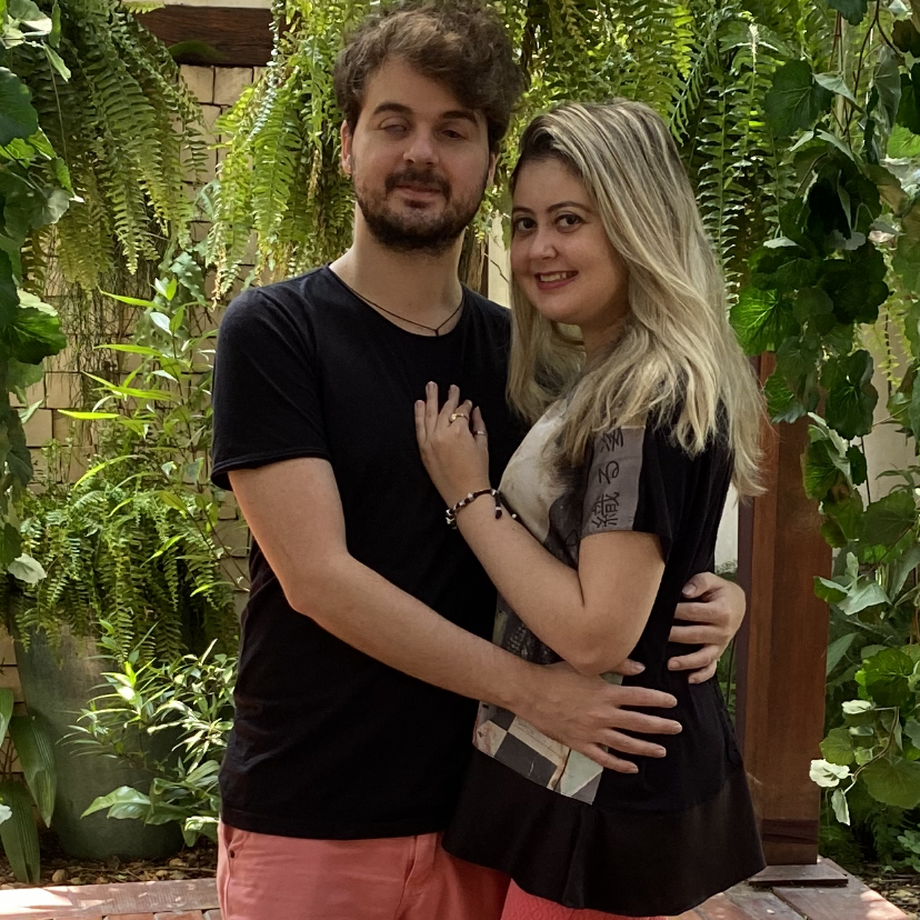 Casal Monique e Rodolfo em um ambiente arborizado, ambos sorrindo e vestindo preto e rosa